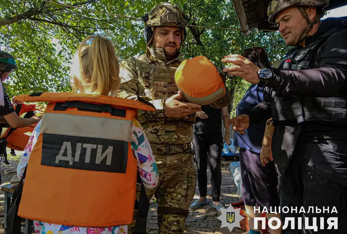 Евакуація дітей із Донеччини. Поліцейські та військові допомагають дитині вдягти захисний шолом, біля них спиною до камери стоїть білява дівчинка в помаранчевому евакуаційному жилеті з написом «ДІТИ». Фото: поліція Донеччини